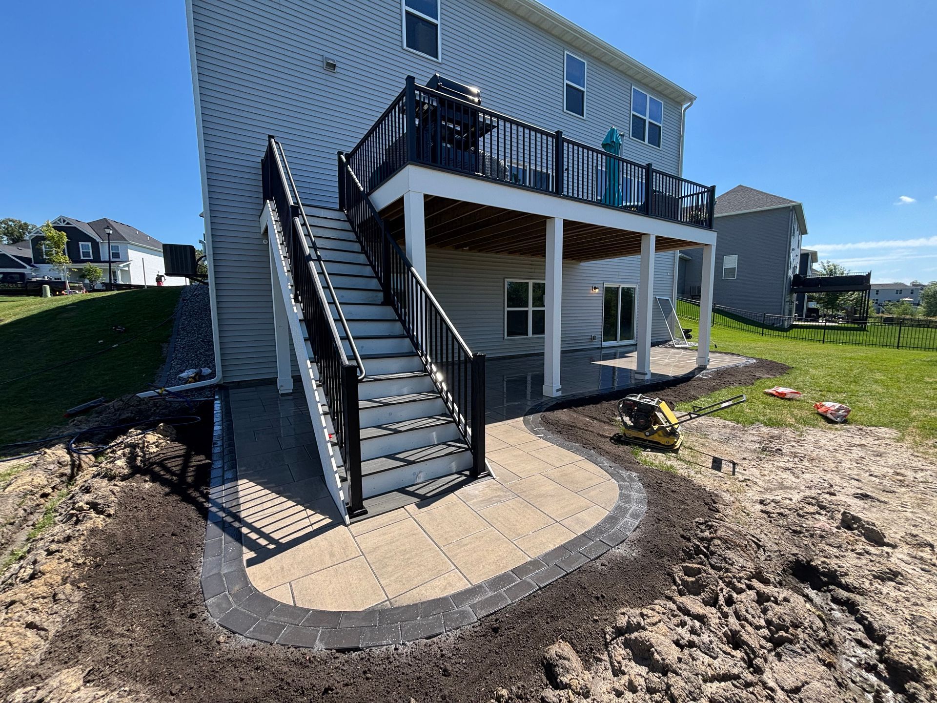 Backyard with a deck, stairs, and a paved patio, surrounded by landscaping with dark soil, blue sky.