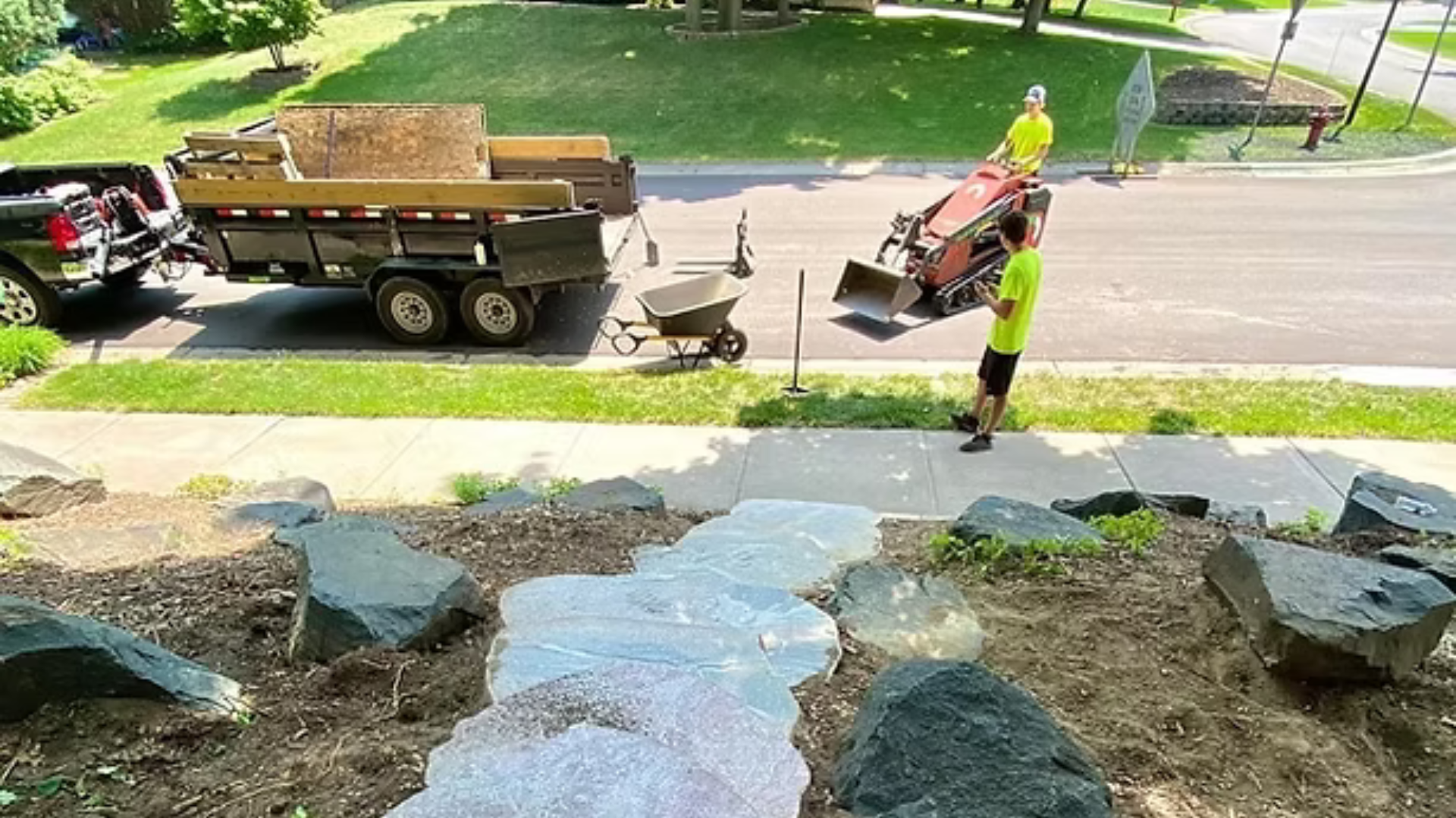 A man is standing on a sidewalk next to a trailer with a wheelbarrow attached to it.