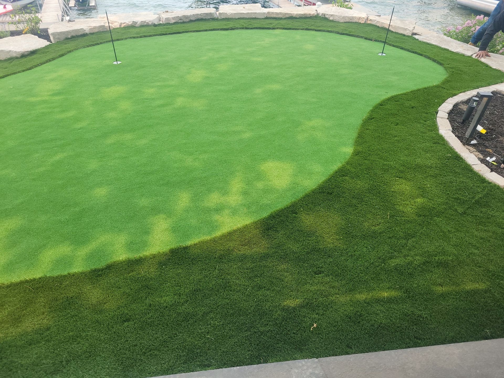 A small artificial turf putting green with darker rough and flags, surrounded by a stone border.