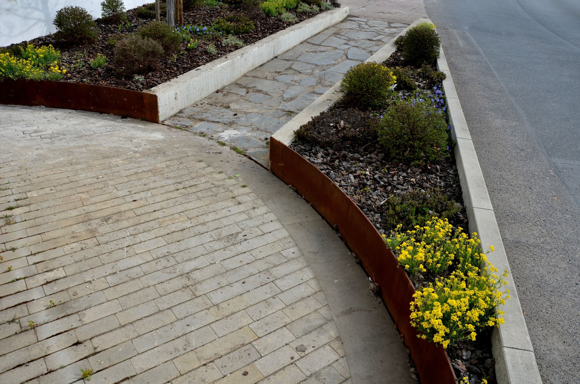 Concrete pathway and raised flowerbeds with rust-colored metal edging and various plants, next to an asphalt road.