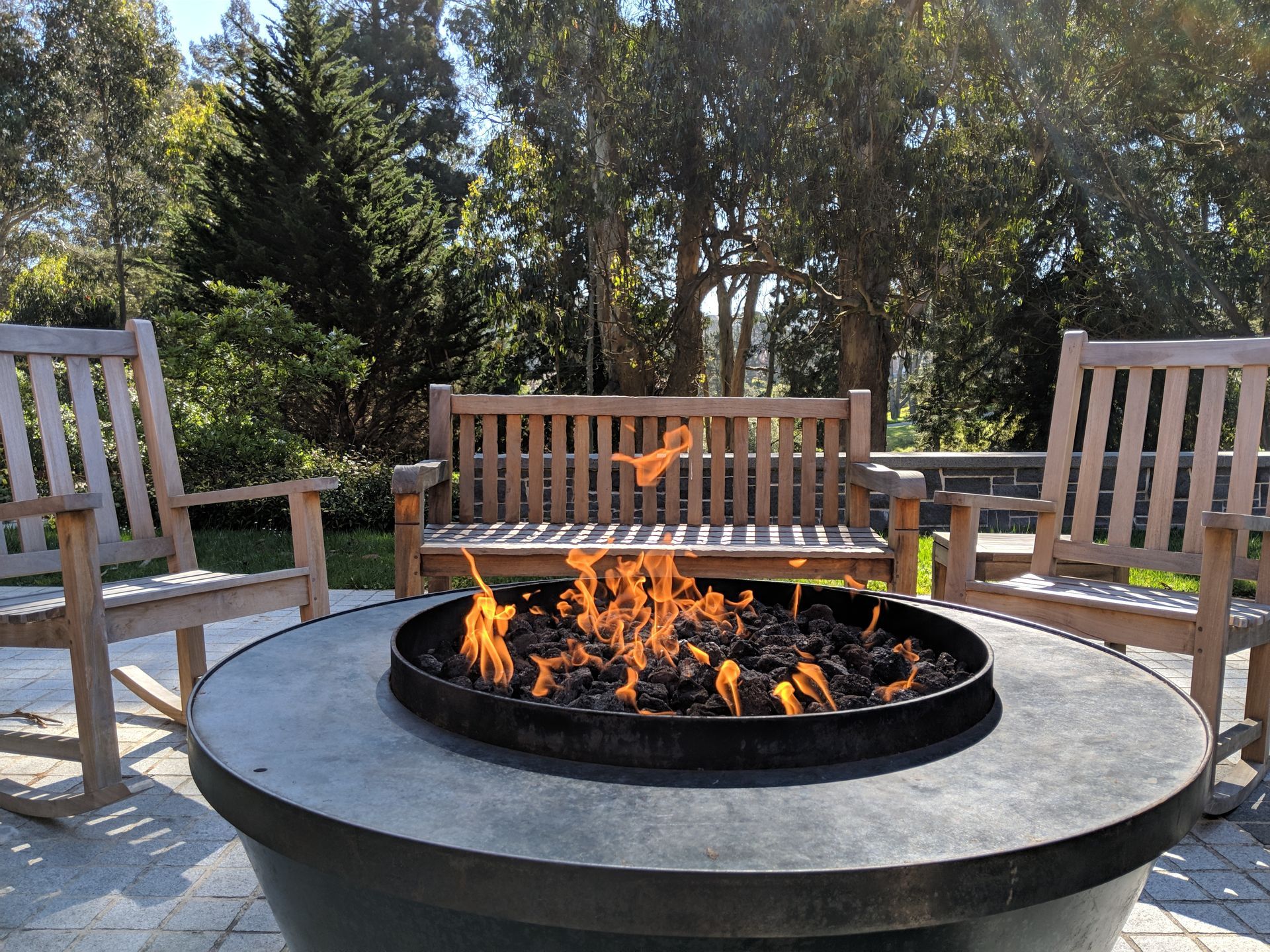 Fire pit with flames, surrounded by wooden chairs and a bench in a sunny outdoor setting.