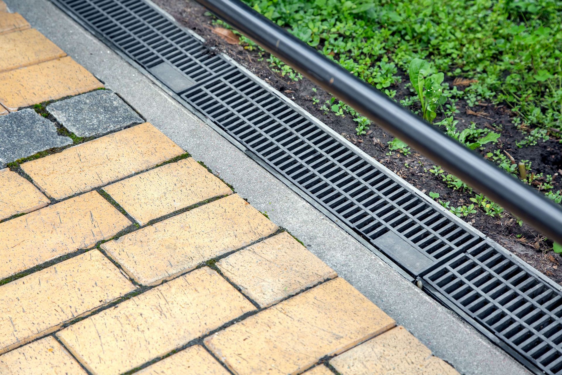 A drain and steel edging on a sidewalk next to a brick walkway.
