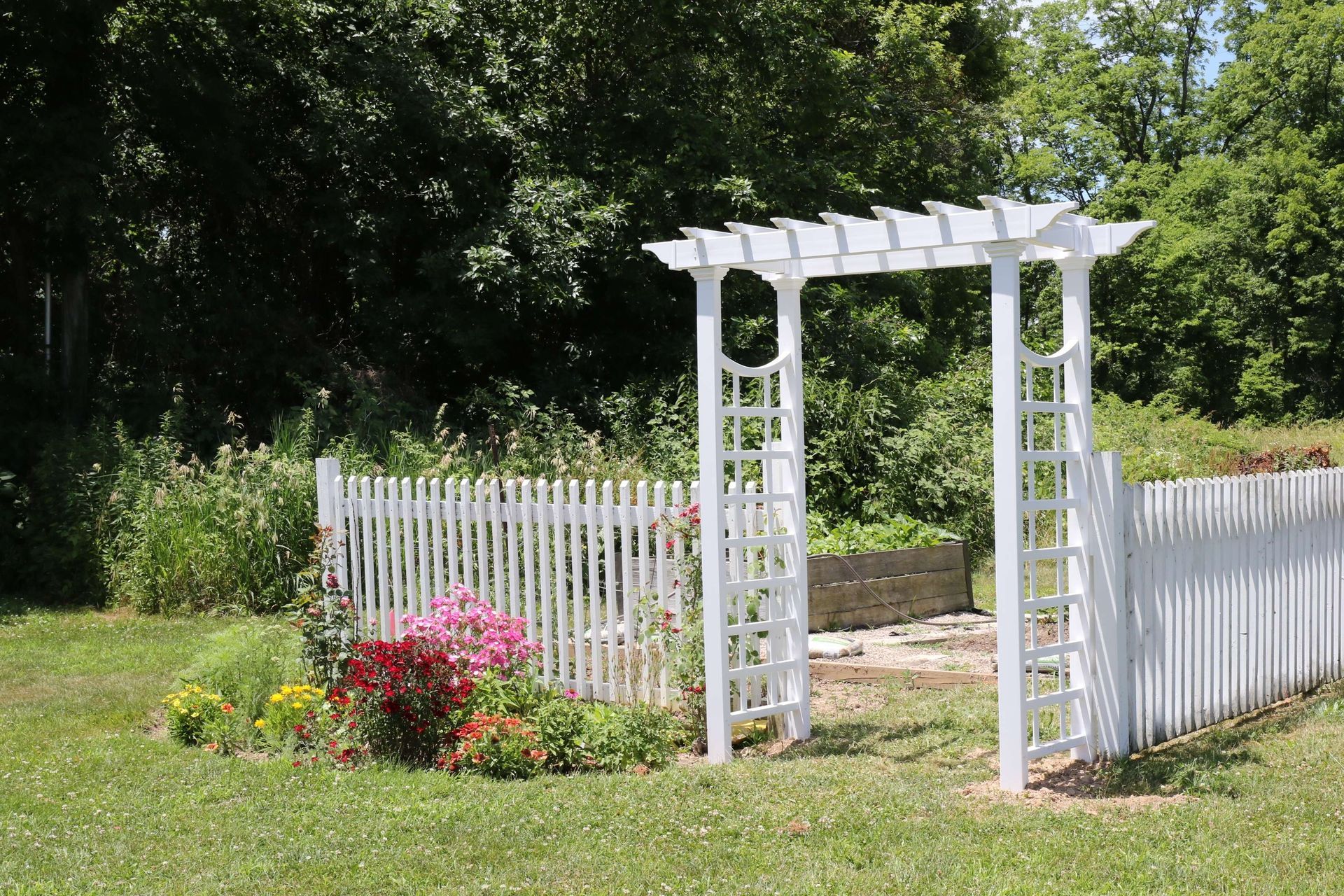 A white arbor is surrounded by a white picket fence in a garden.
