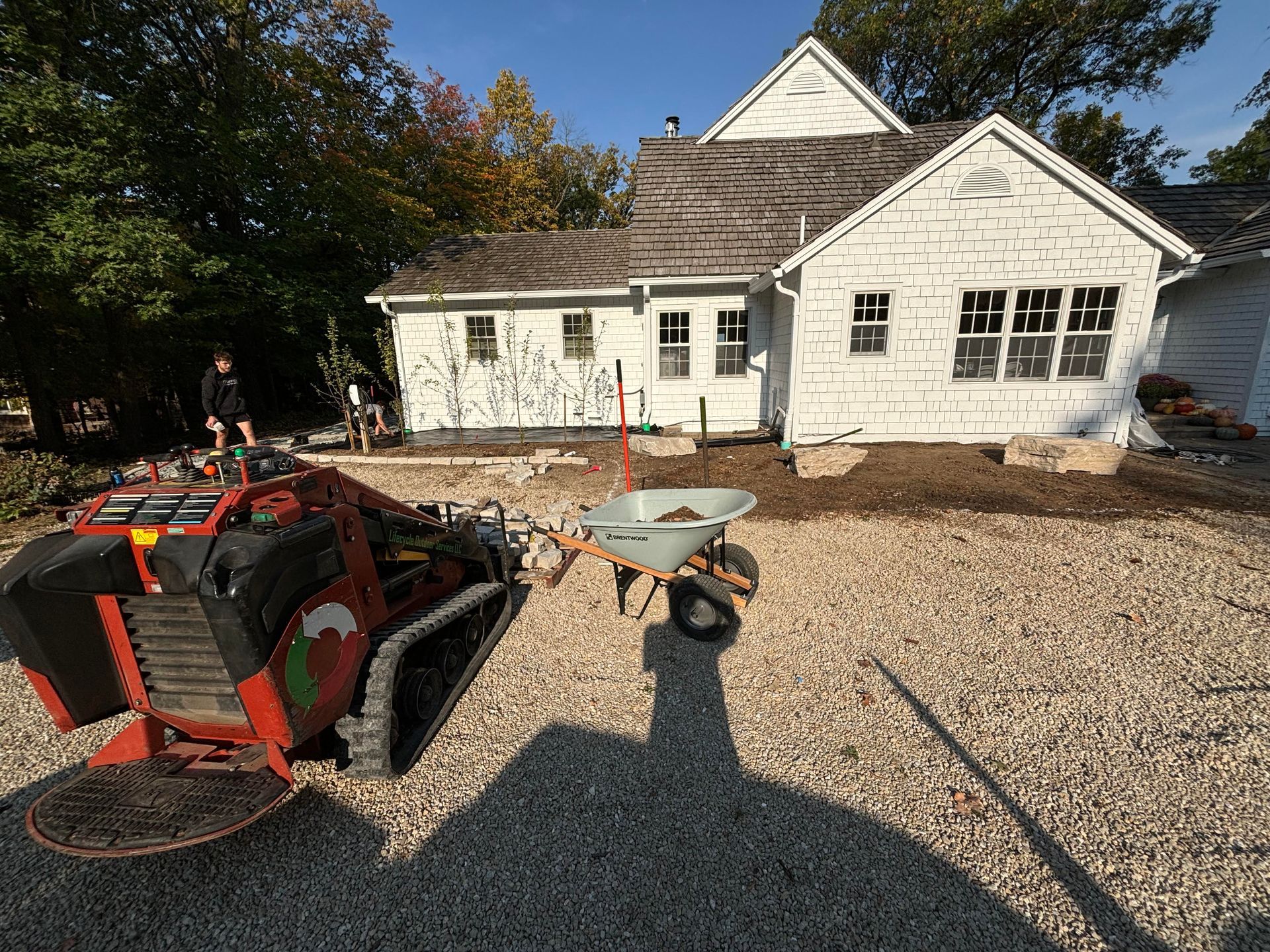 A skid steer pulling a wheelbarrow, working near a white house. Sunlight casts shadows.