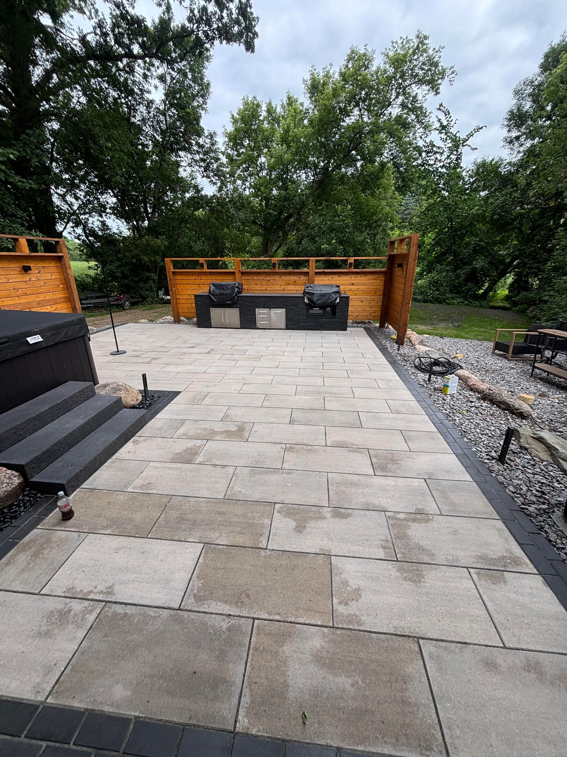 A patio with grey stone pavers and a built-in grilling area, set against a natural backdrop of trees.