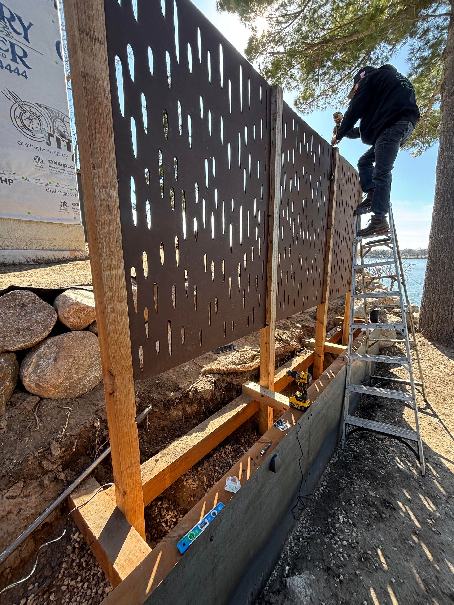 Man on ladder installing a decorative metal fence near water. Brown panels, wooden posts, sunny day.