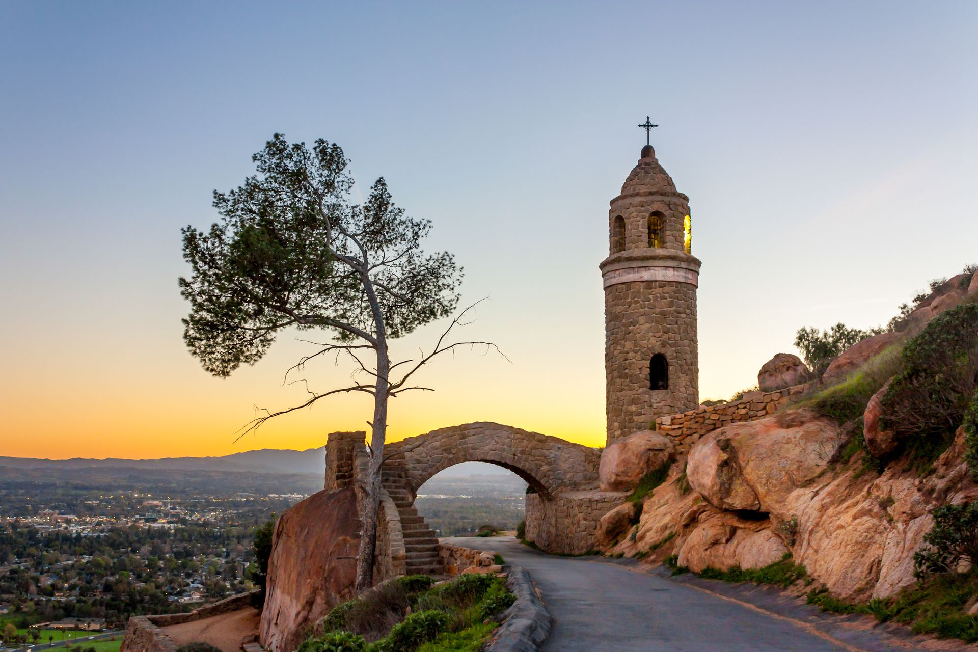 Stone tower and arch bridge on a hillside road at sunset, overlooking a city.