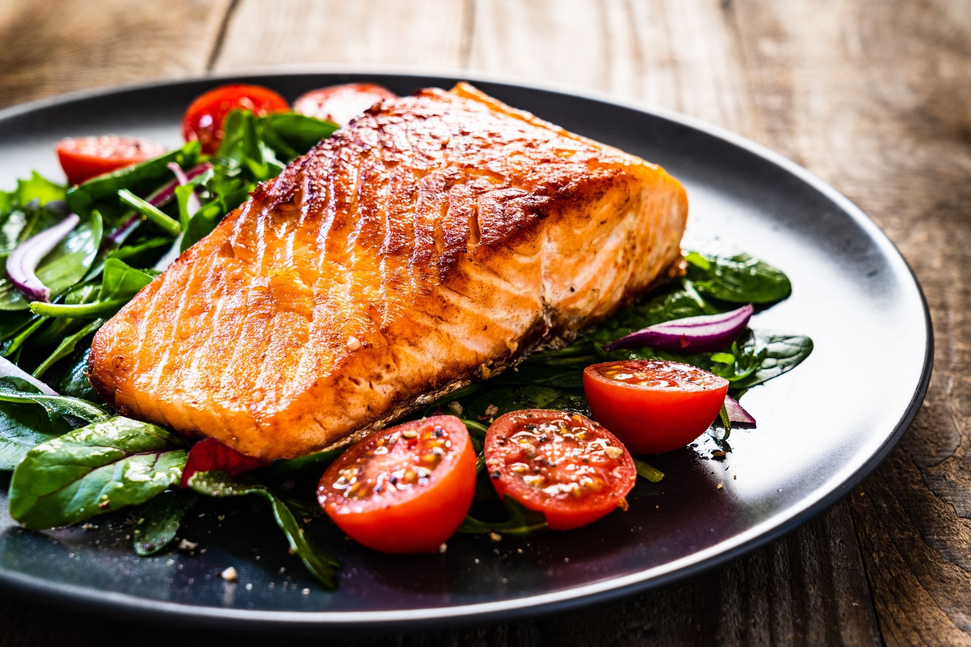 Seared salmon fillet with salad, cherry tomatoes, and red onion on a dark plate, on a wooden surface.