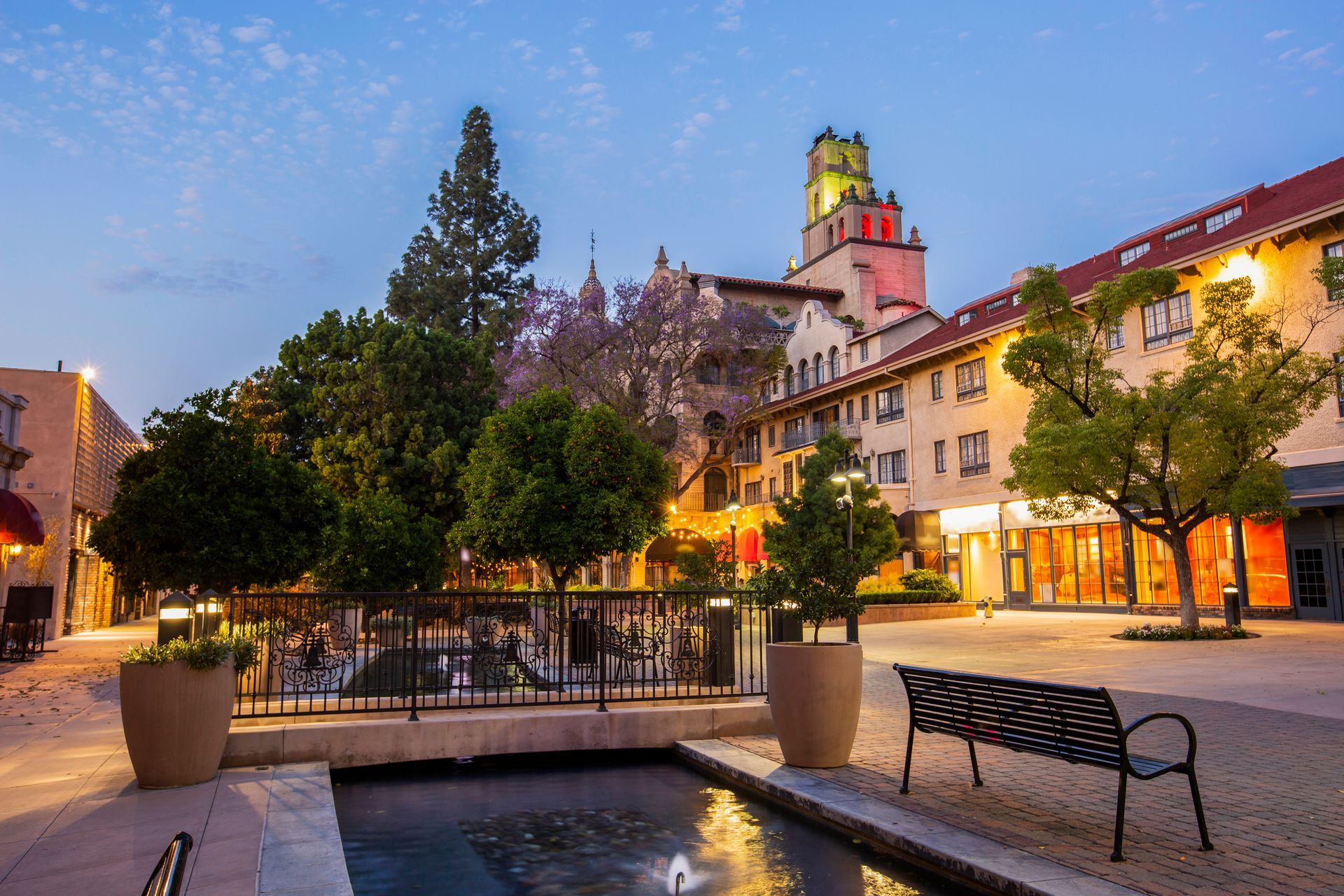 A European-style plaza with shops, trees, and a fountain at dusk; warm lighting.