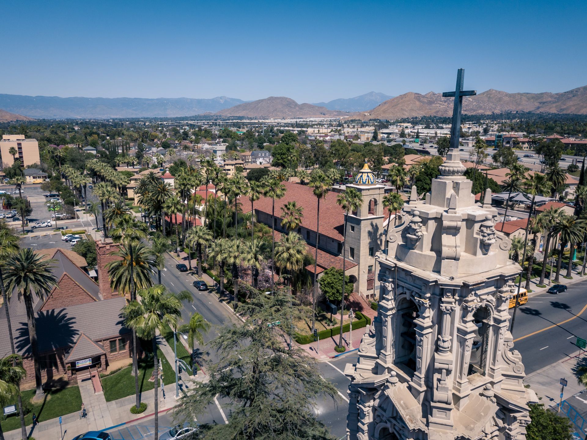 Aerial view of a church with a tall cross, palm trees, and a cityscape in the background.