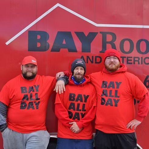 Three people in red shirts and hats pose in front of a red sign for Bay Roofing.