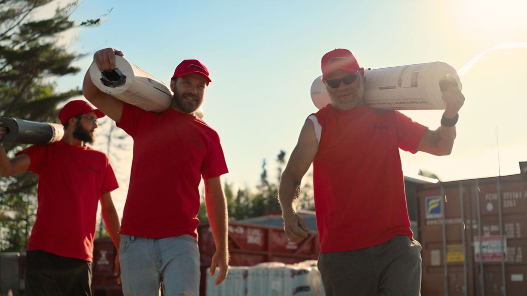 Three people in red shirts and hats carrying objects on their shoulders outdoors.