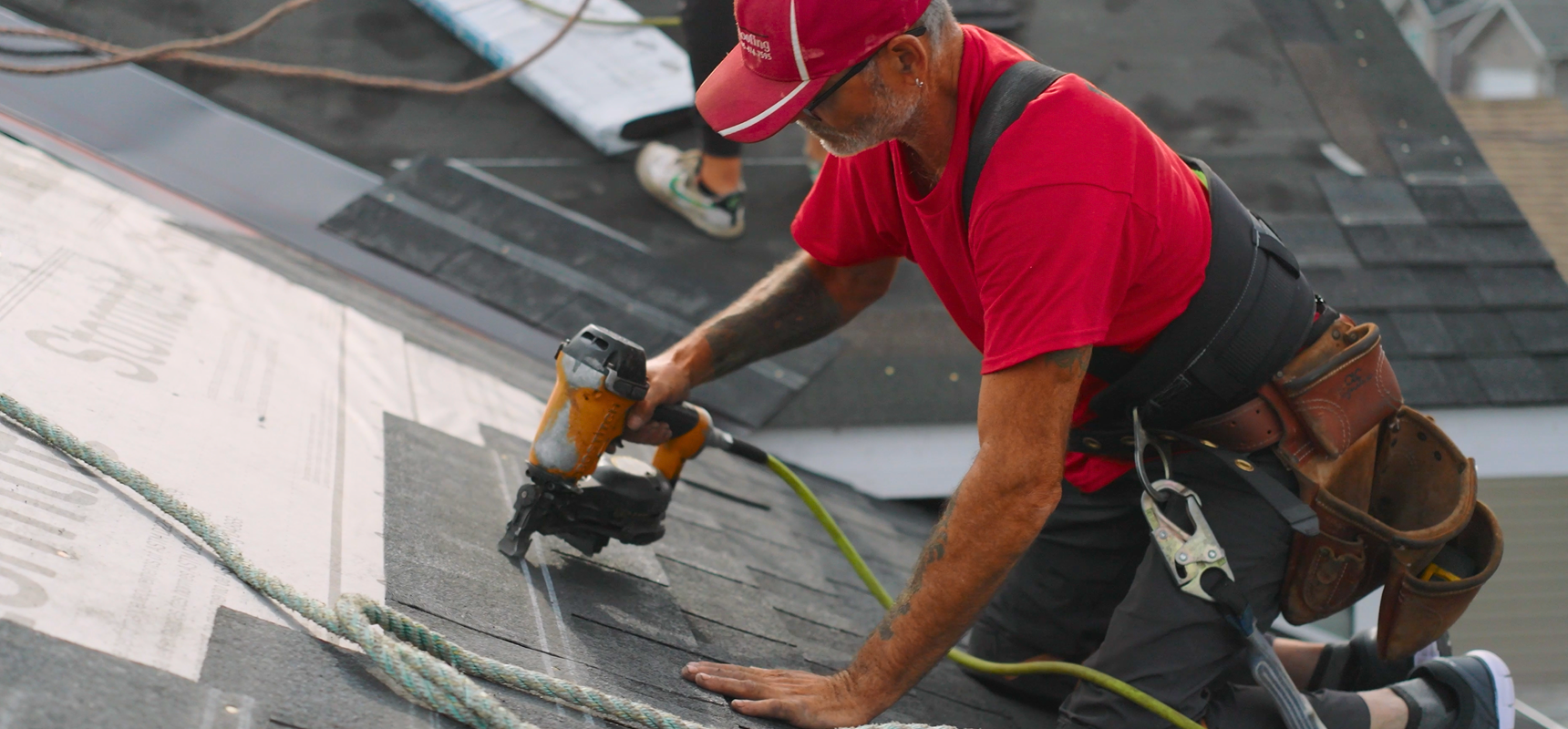 Roofer in a red shirt and cap using a nail gun on a rooftop, with another person visible in the background.