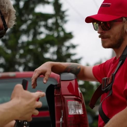 Man in red shirt and cap leans on truck, talking to another person.