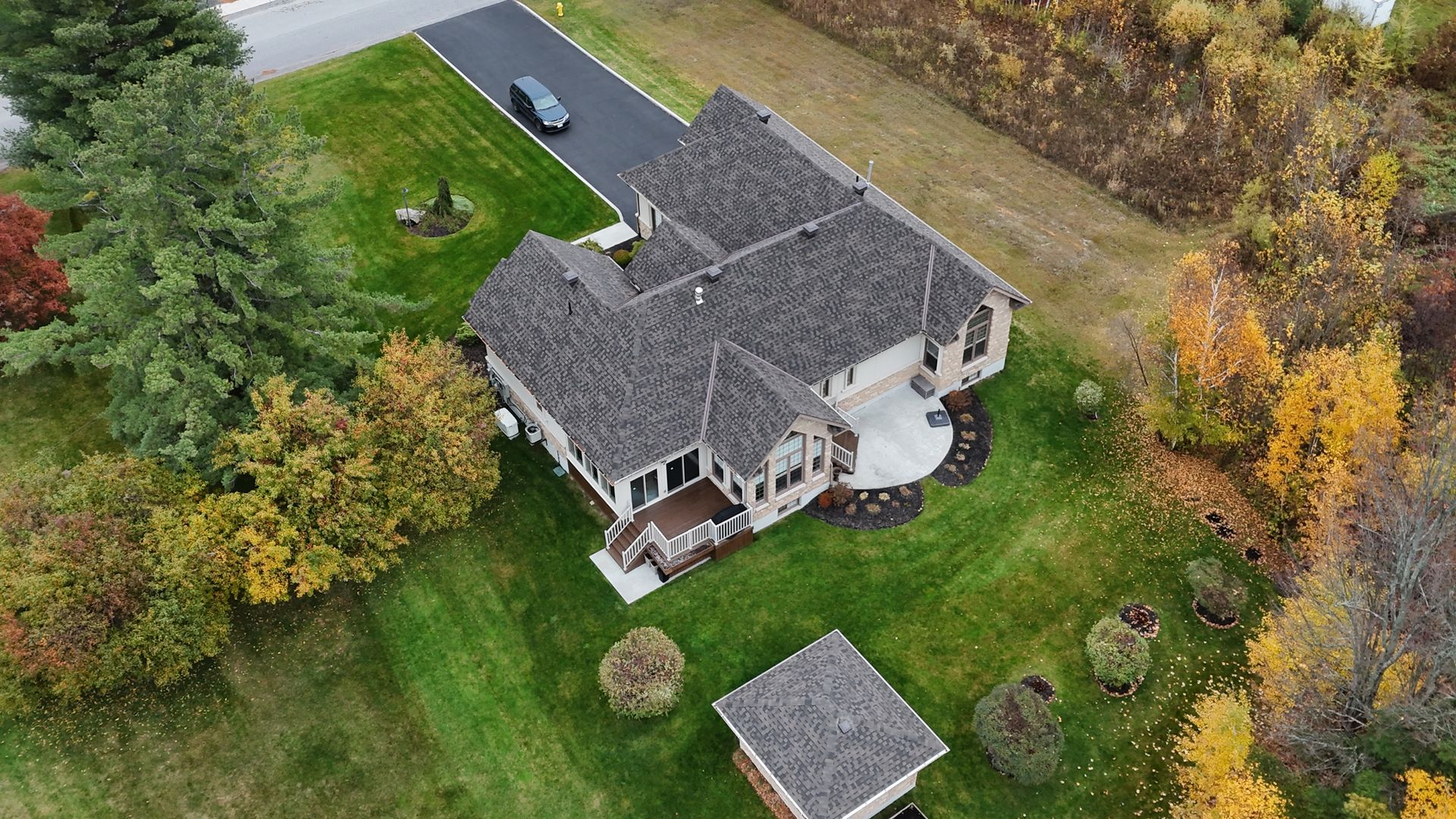 Aerial view of a house with a dark roof and paved driveway, surrounded by green grass and fall foliage.