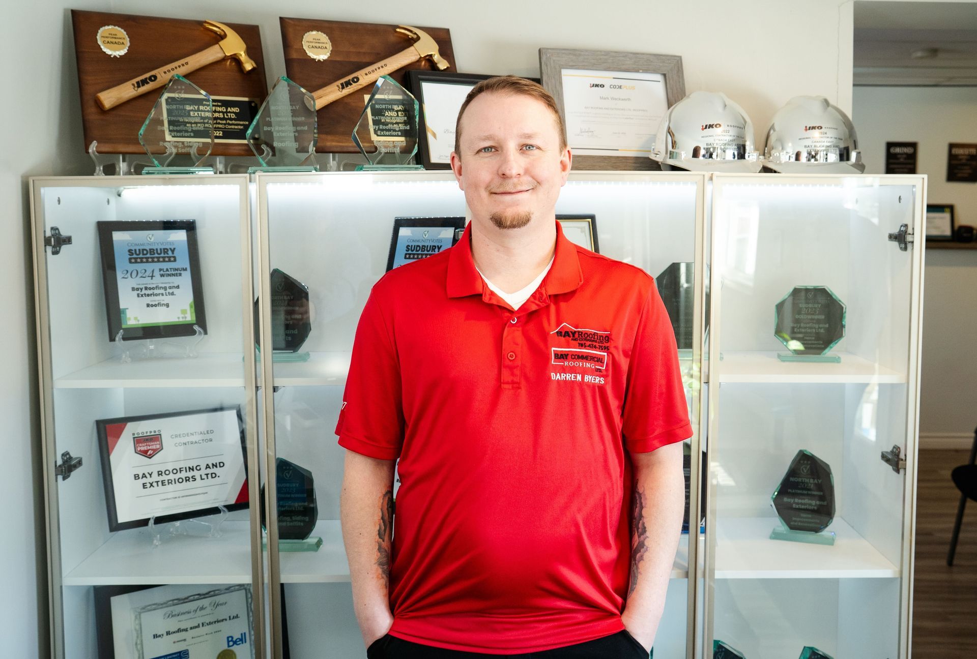 Man in red shirt stands in front of a cabinet displaying awards and hard hats.
