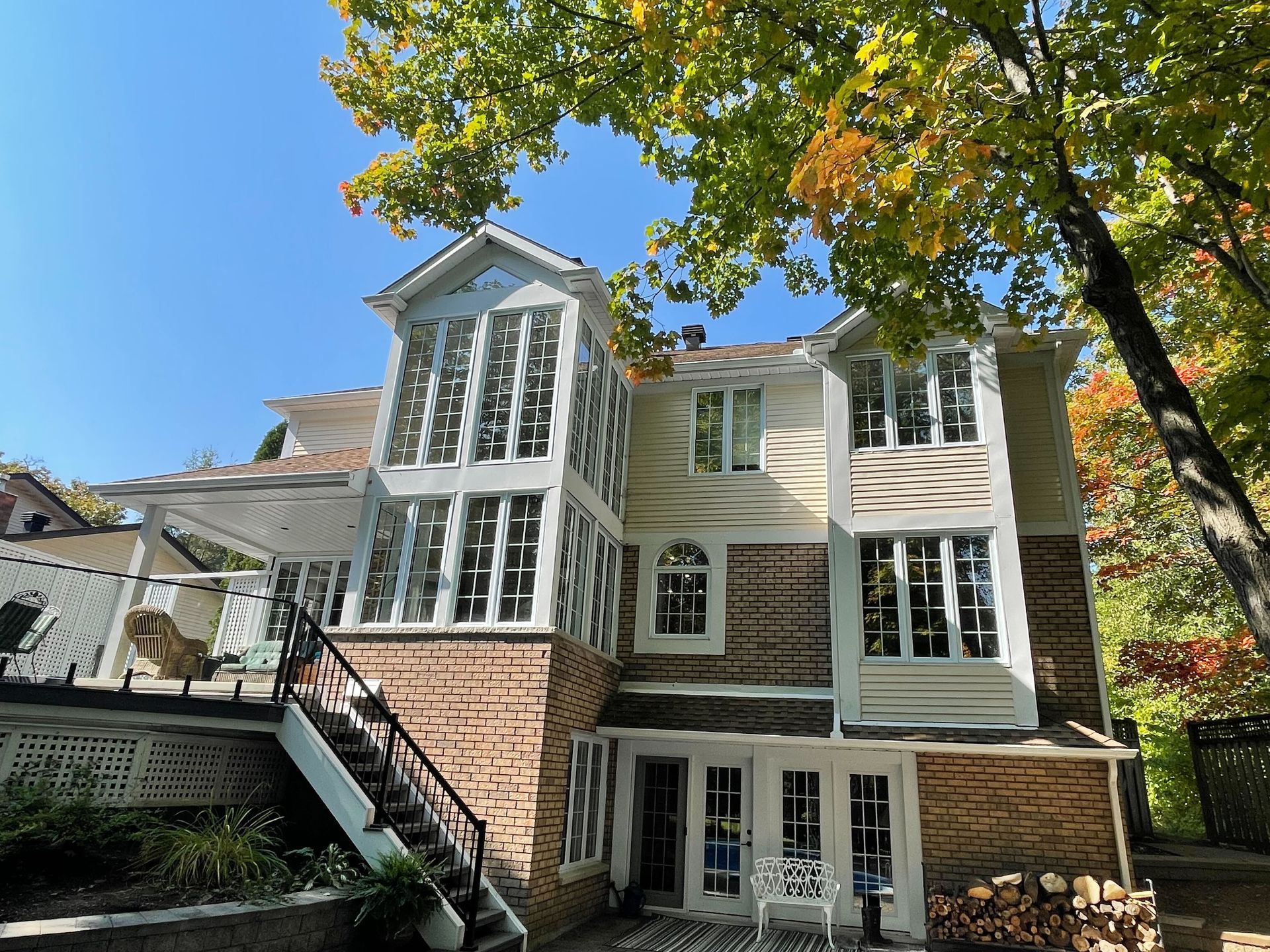 Multi-story house with brick and white siding, many windows, and a staircase leading to a deck; autumn foliage.