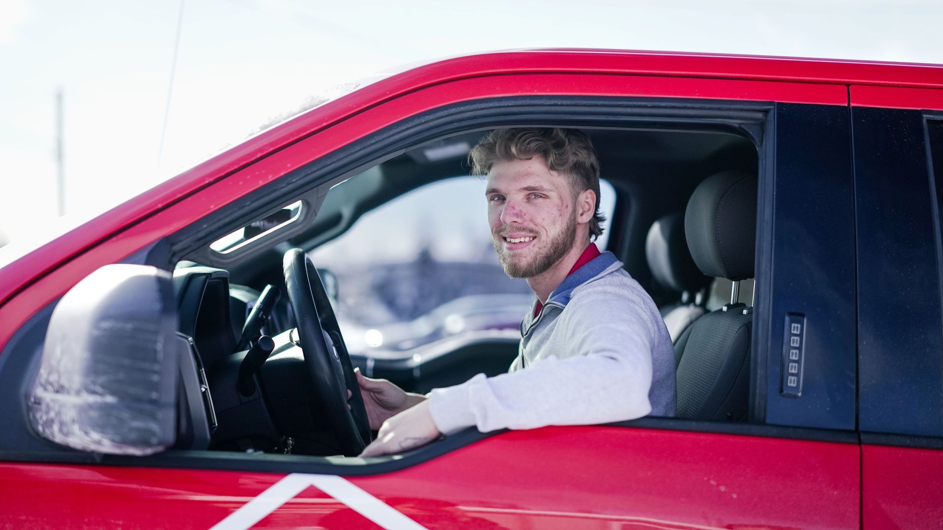 Man smiling in a red pickup truck, sunny outdoor setting.