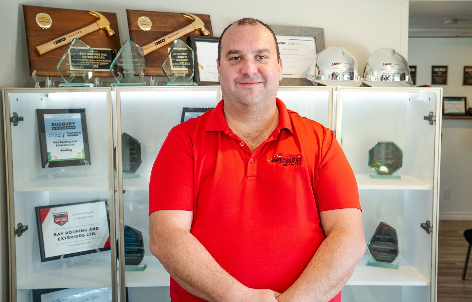 Man in red shirt smiling in front of a display case with awards and helmets.