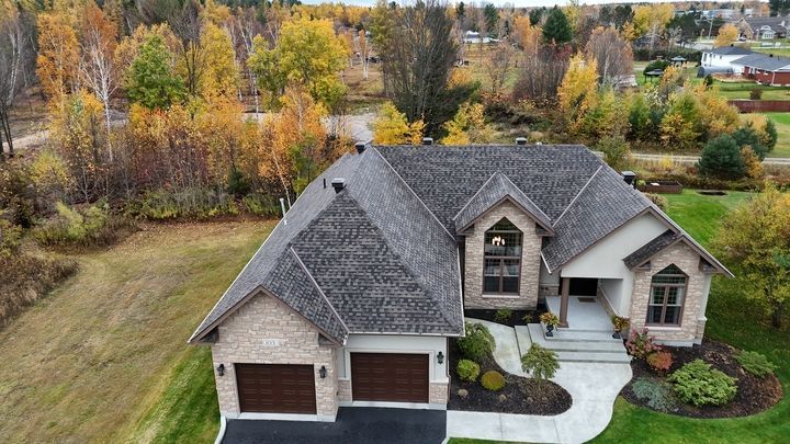 House with a gray roof and tan brick facade surrounded by fall foliage. Two-car garage, manicured lawn.