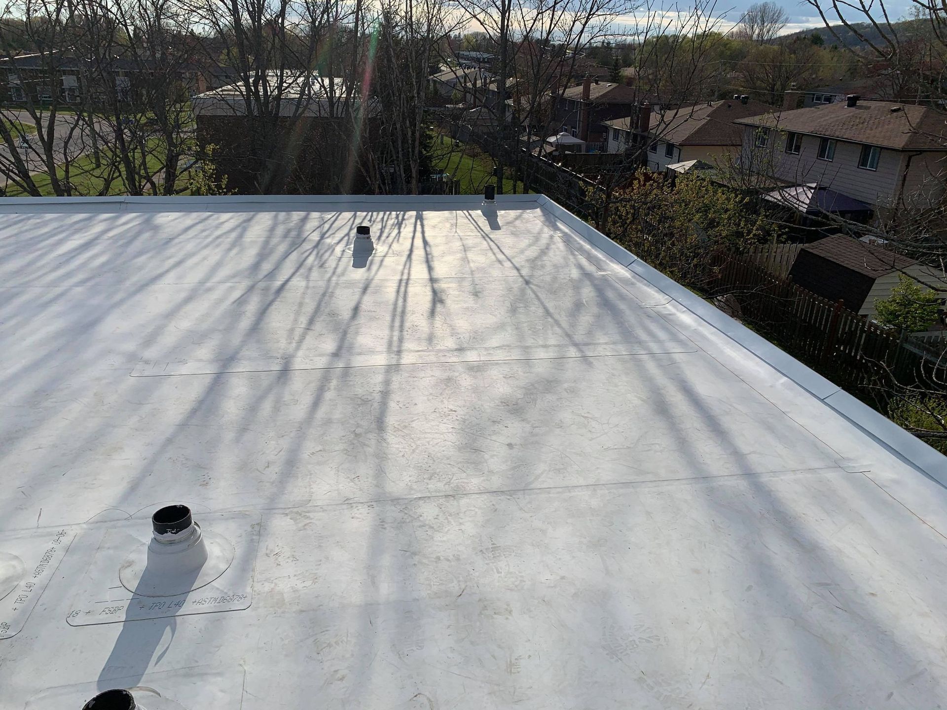 Flat, white commercial roof with vent pipes, viewed from above, with trees and buildings in the background on a sunny day.