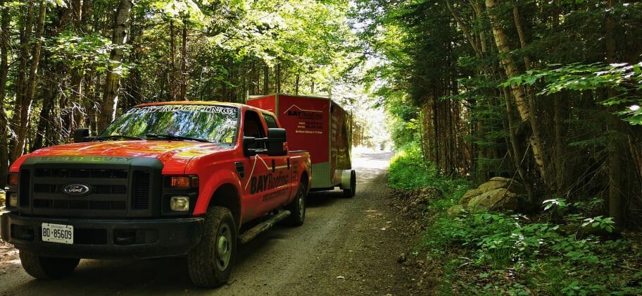 Red pickup truck towing a red trailer on a dirt road through a forest.