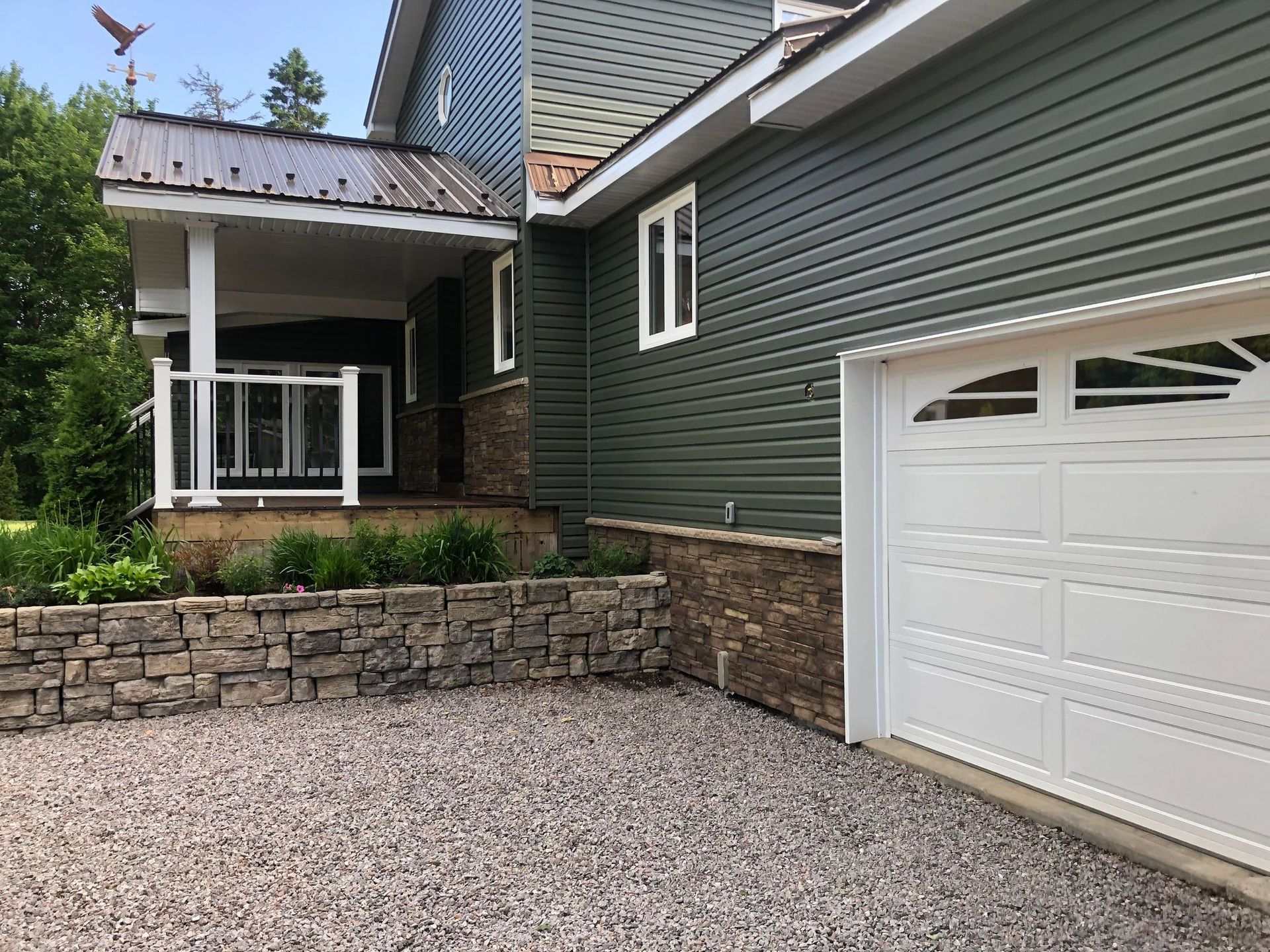 Green-sided house with a white garage door, stone accents, and a gravel driveway.