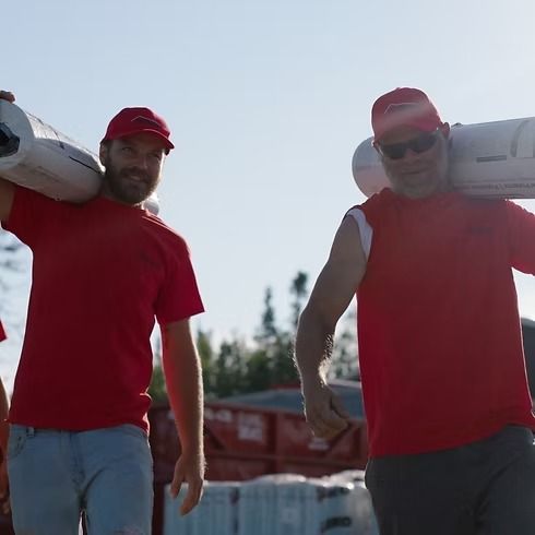 Two men in red shirts and hats carrying white objects on their shoulders outdoors.