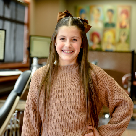 A smiling child with braces, wearing a brown sweater and matching bow in their hair, stands in an office setting.