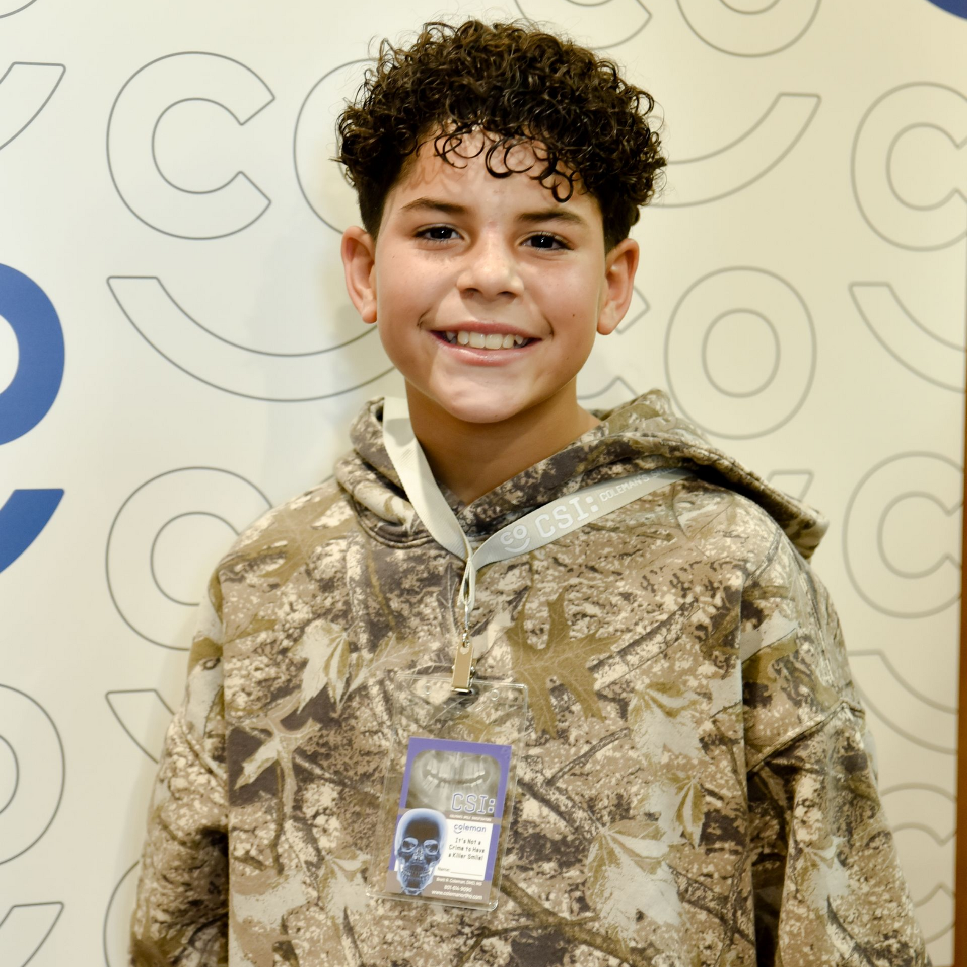 Young person with curly hair smiles, wearing a camo hoodie, in front of a patterned wall.