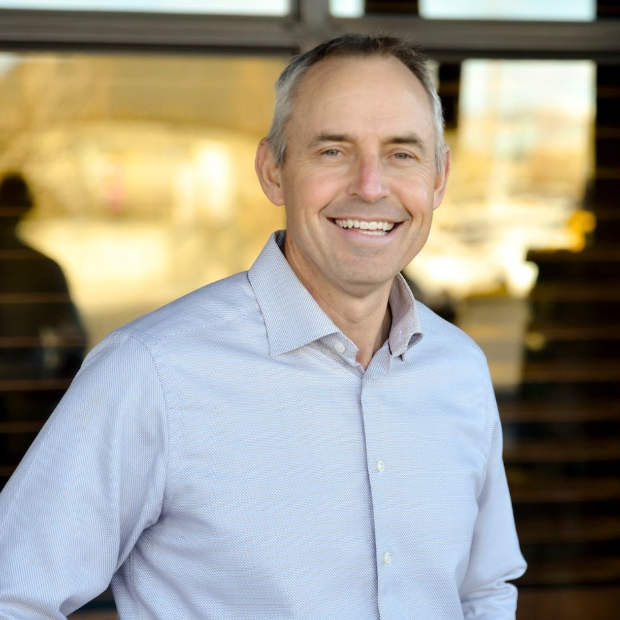 Man smiling, wearing a light blue button-down shirt, standing in front of a building.