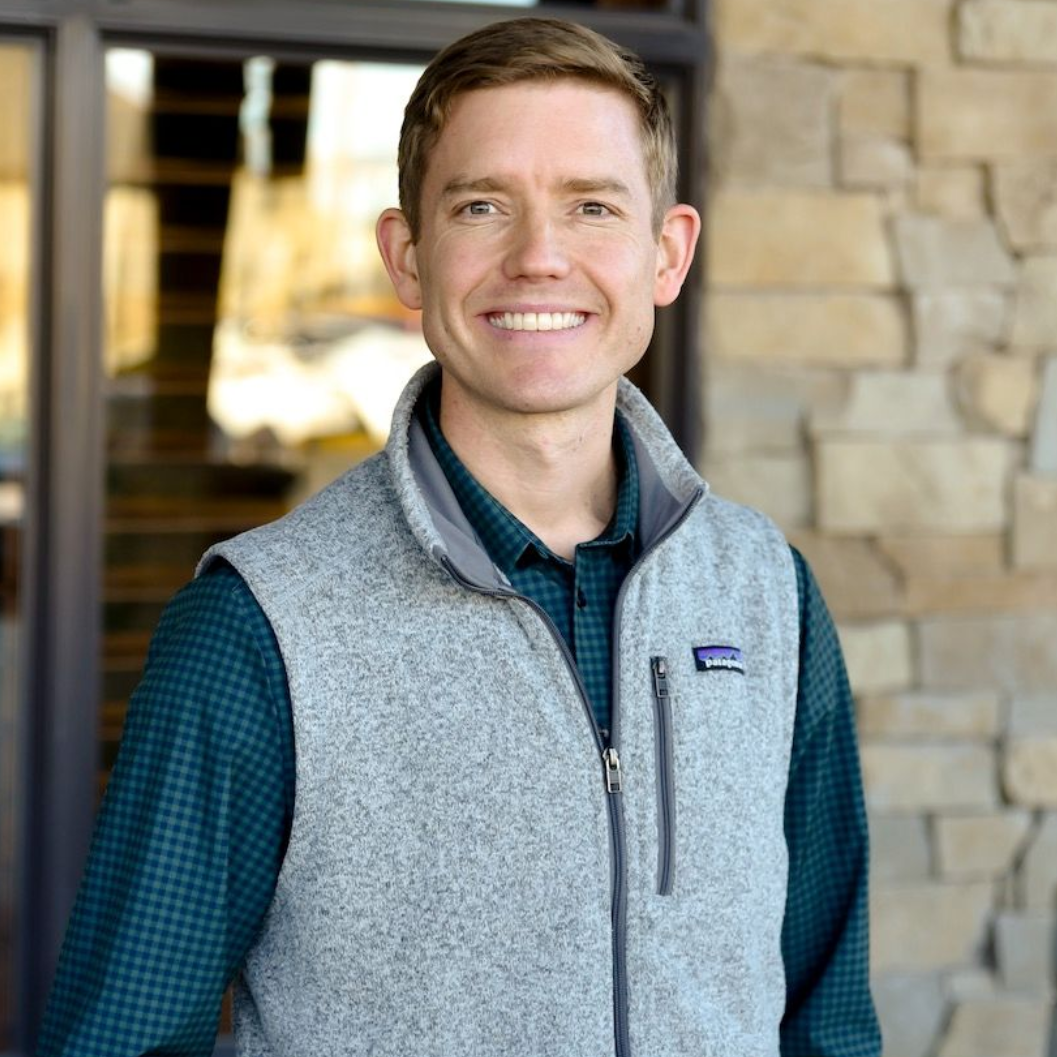 Man in gray vest smiles, standing outside a building with stone and glass elements.