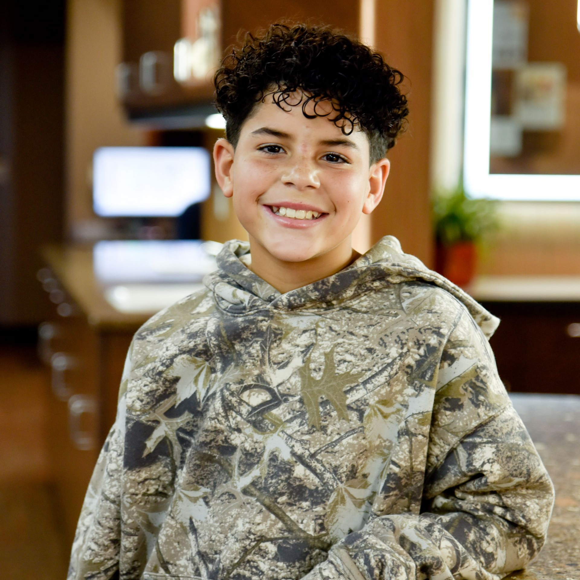 A smiling person with curly dark hair wearing a camouflage hoodie in a brightly lit indoor space.
