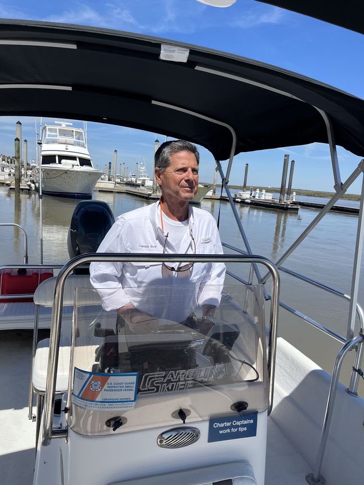 A person wearing a white shirt pilots a small boat in a marina with other boats docked in the background.