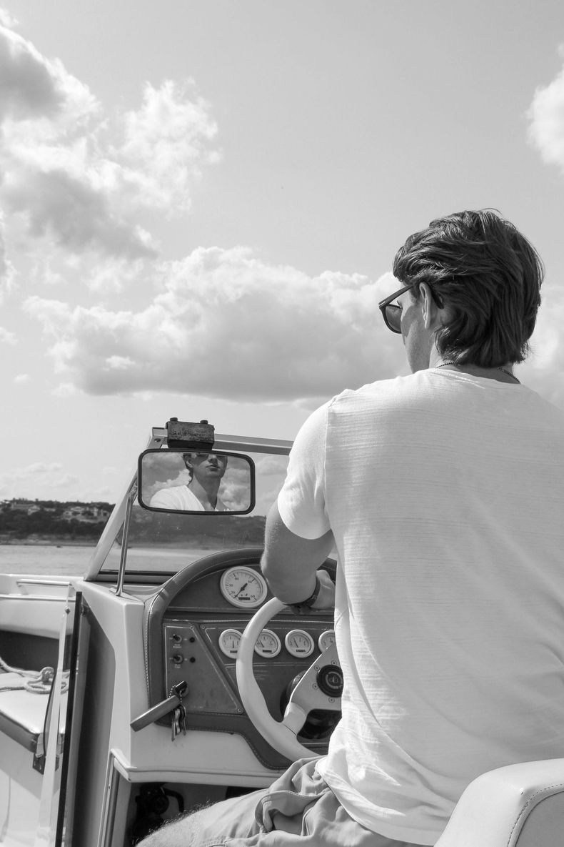 A person in a white t-shirt and sunglasses steering a motorboat on a sunny day, seen from behind.