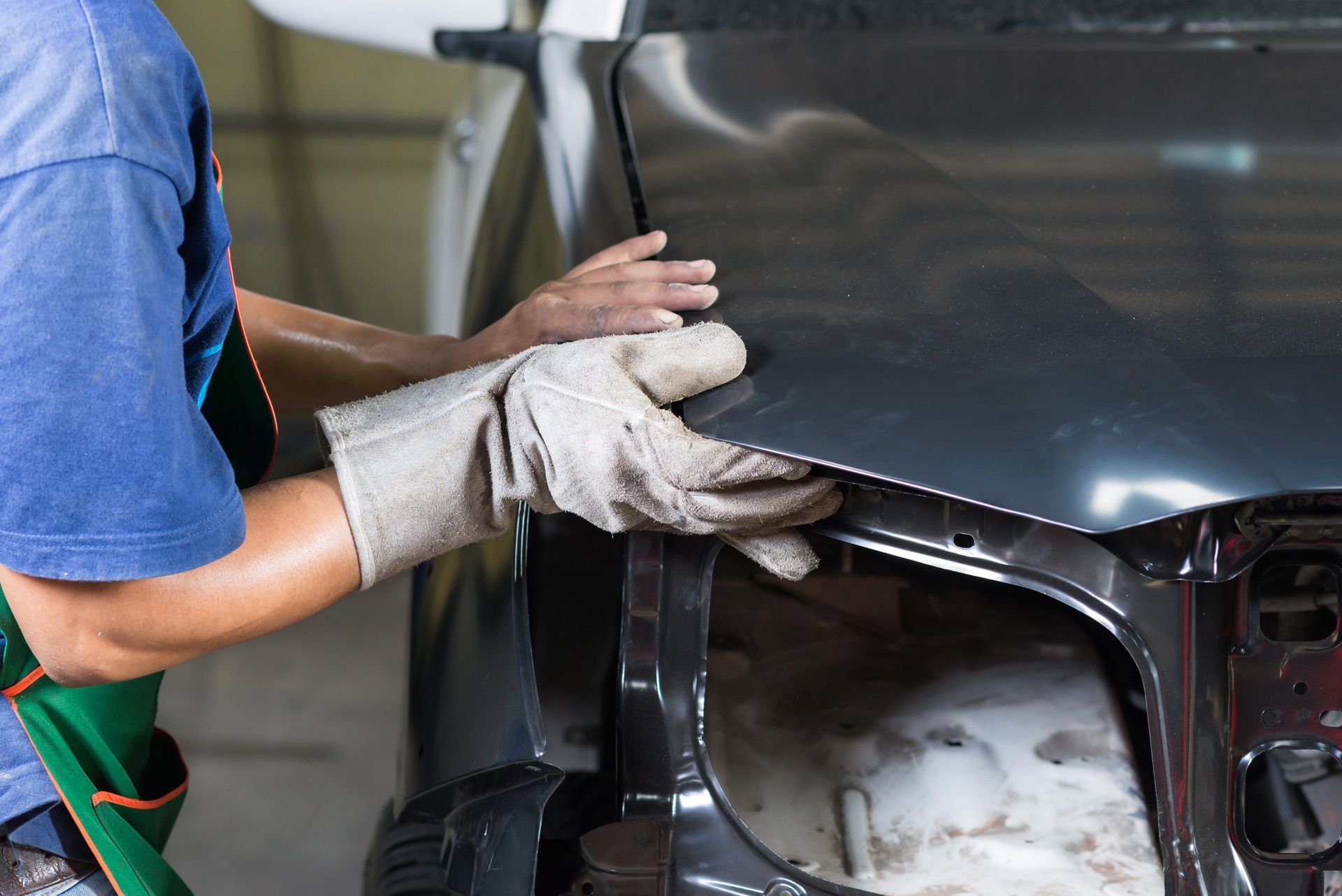 A person wearing work gloves aligns a dark car hood onto the vehicle frame in an auto body shop.