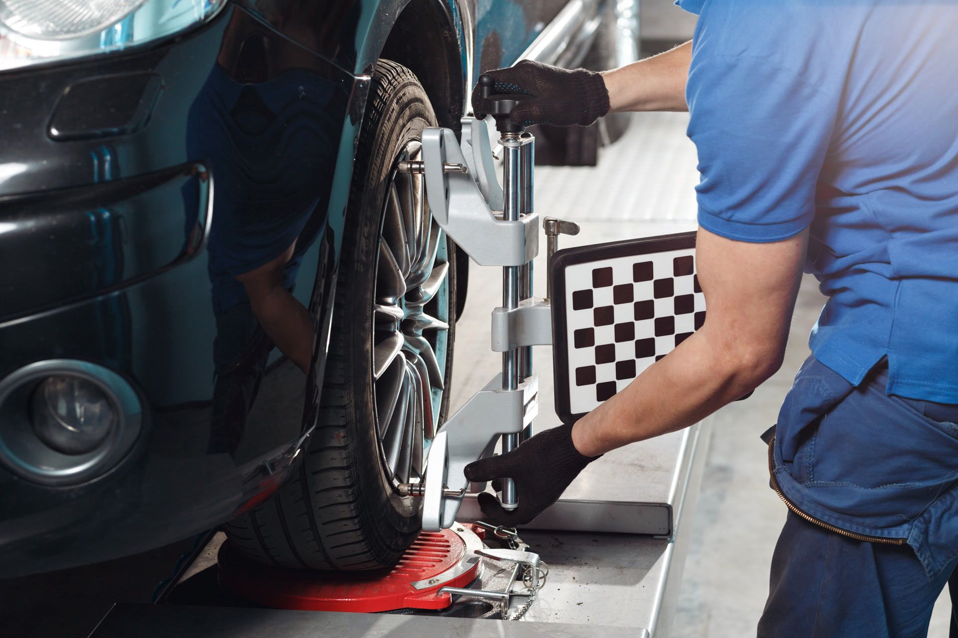 A technician in a blue uniform and black gloves adjusts a wheel alignment sensor onto a car tire in an auto shop.