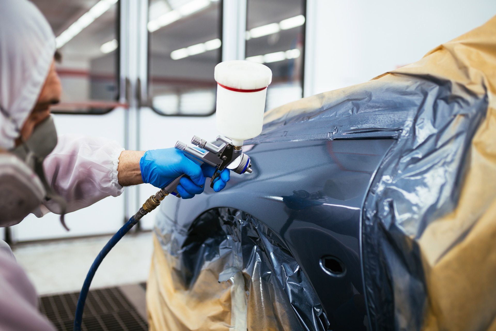 An auto body technician in protective gear uses a spray gun to paint the dark gray body of a car in a shop.