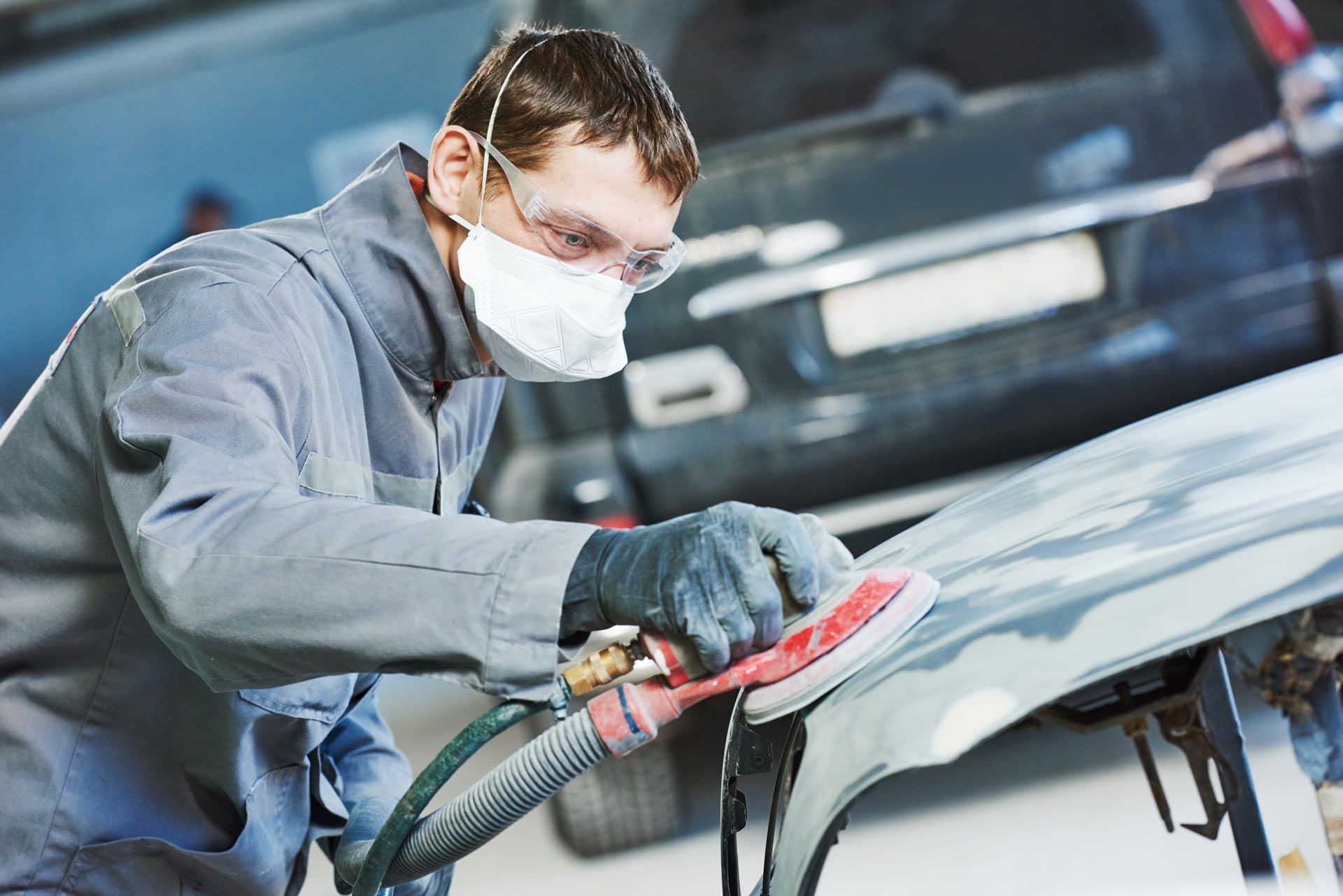 A worker wearing protective gear uses a sander on a car body panel in a repair shop.