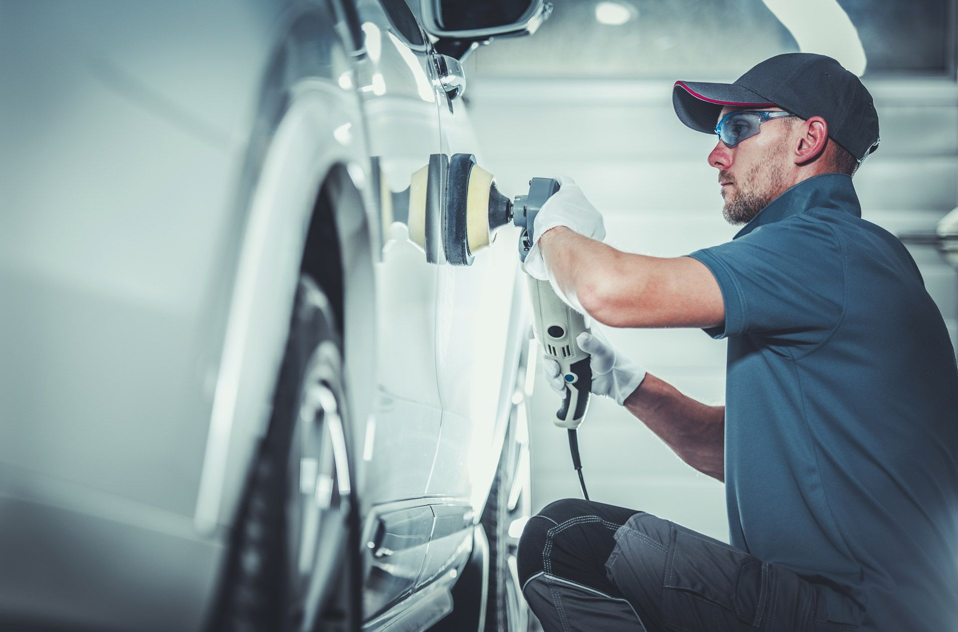 A person wearing a cap, glasses, and gloves uses a power buffer to polish the side of a white car in a garage.