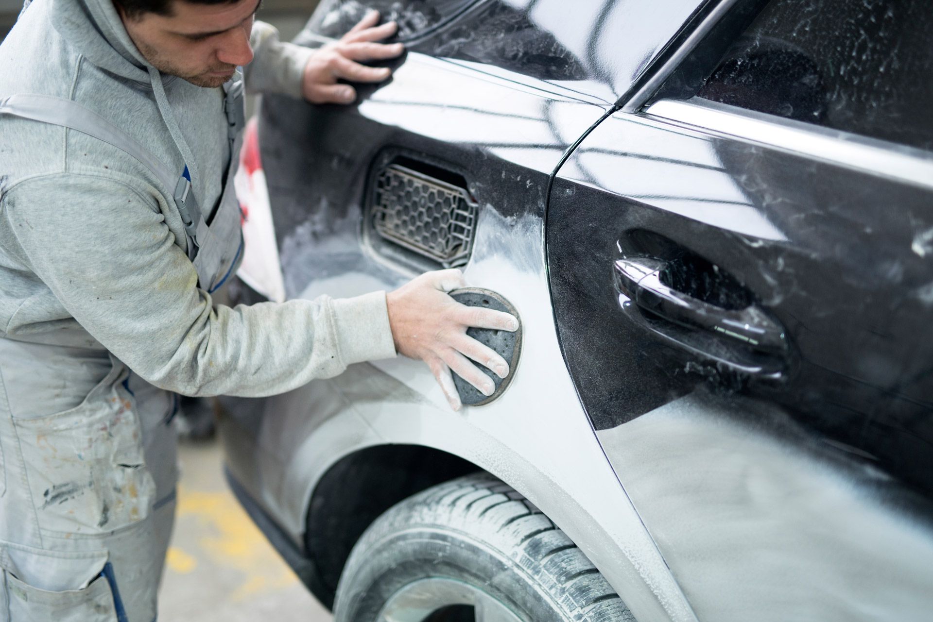 A person in work coveralls sands the gray, prepped rear quarter panel of a car in an auto body repair shop.