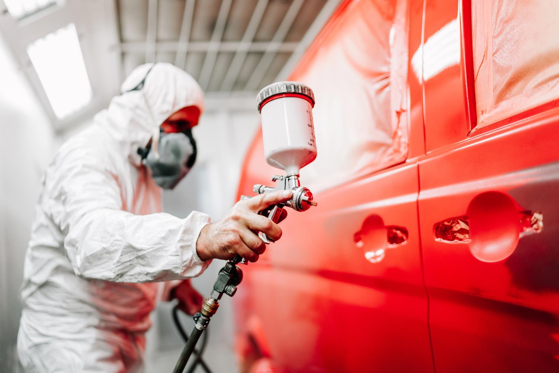 A person in protective gear uses a spray gun to paint the side of a bright red vehicle in a professional workshop.