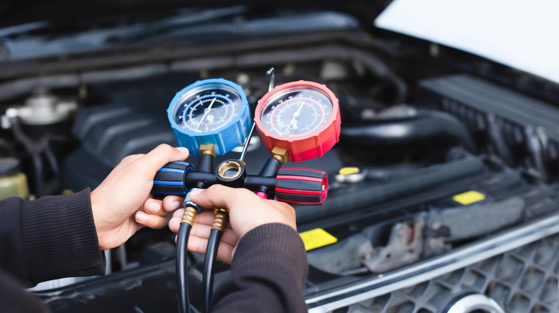 A person holds an automotive AC manifold gauge set in front of an open car hood.