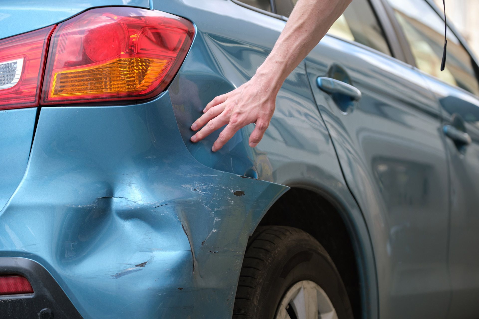 A hand touches a deep dent and paint scrape on the rear quarter panel of a light blue car.