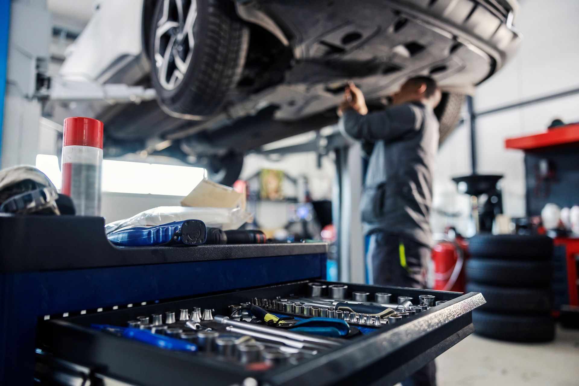 A mechanic works on the undercarriage of a car lifted in a garage, with a foreground workbench holding tools.
