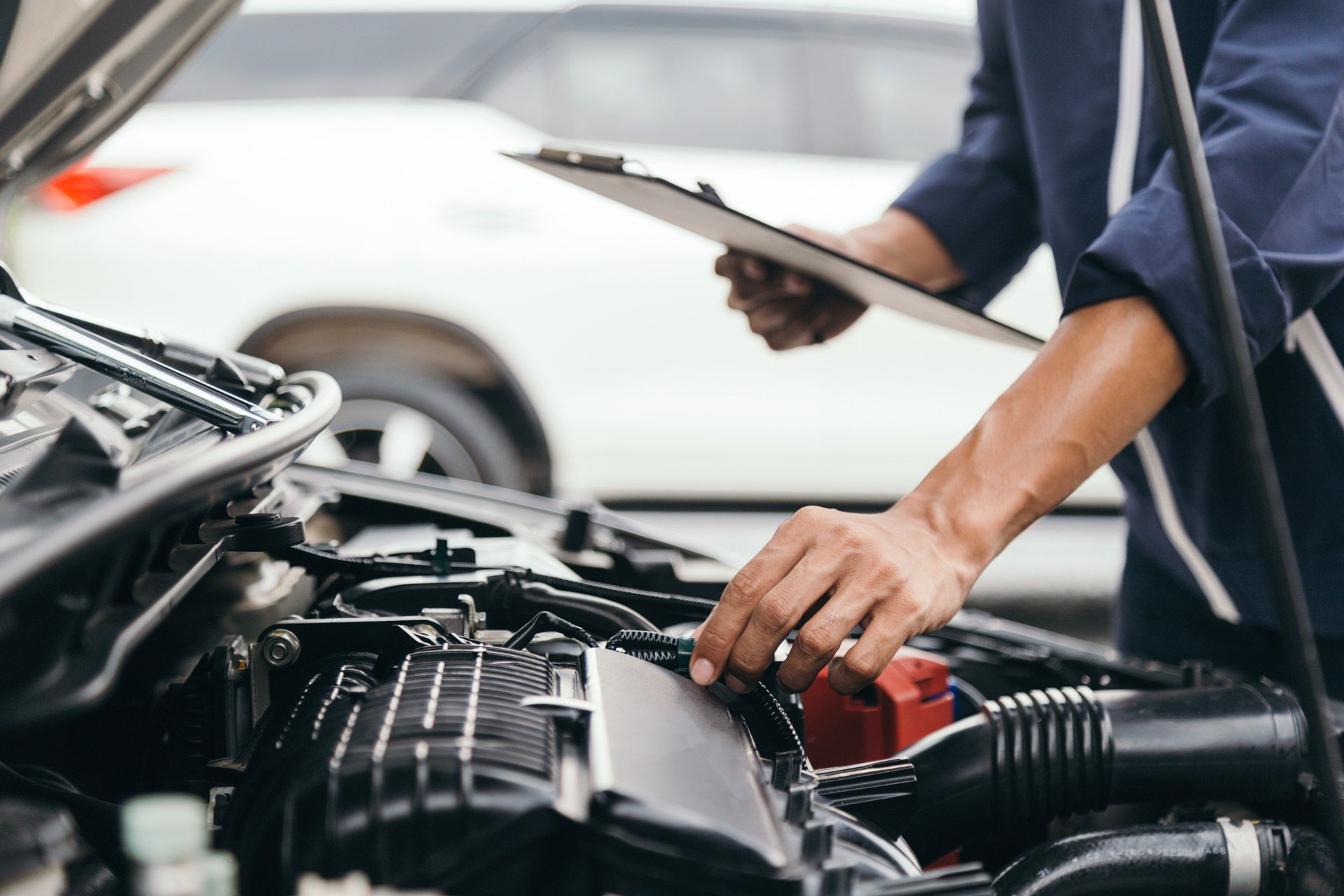 A mechanic in a blue uniform inspecting an open car engine while holding a clipboard, with a blurry car in the background.