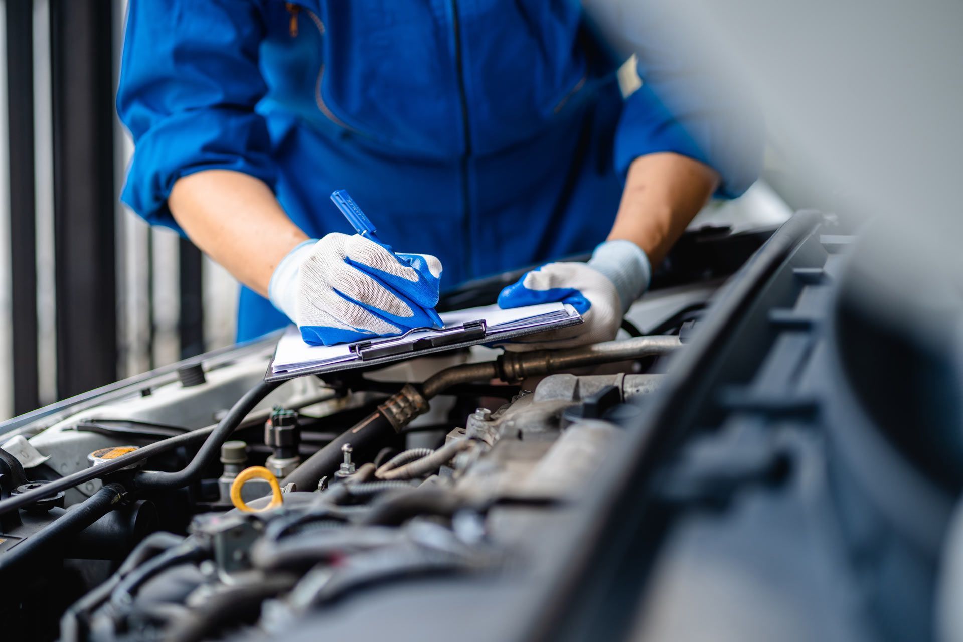 A mechanic in a blue uniform and gloves writes on a clipboard while inspecting an open car engine.