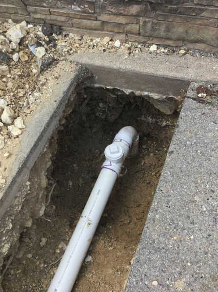 A trench dug in concrete reveals a white PVC pipe with a valve near a brick wall.