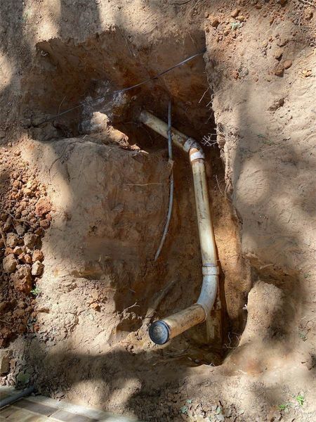 Pipes exposed in a dirt trench; a section of white PVC plumbing with electrical wires visible.