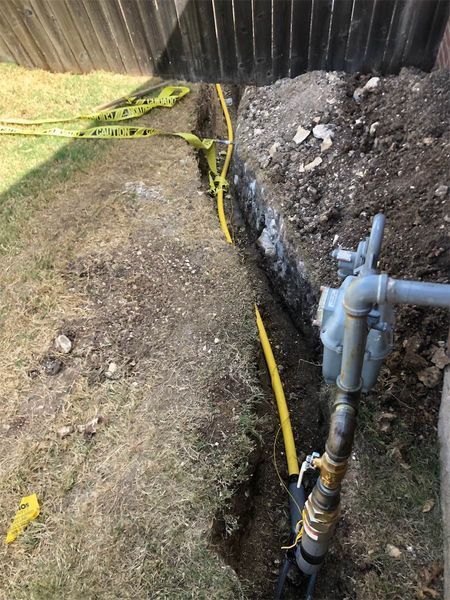 Trench in yard with yellow gas line exposed, caution tape, near fence and gas meter.